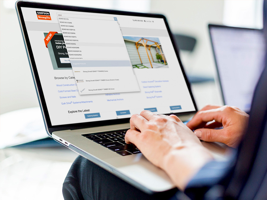 Image of laptop resting on a man's lap. He is typing and the screen is displaying the search feature on the Simpson Strong Tie website.