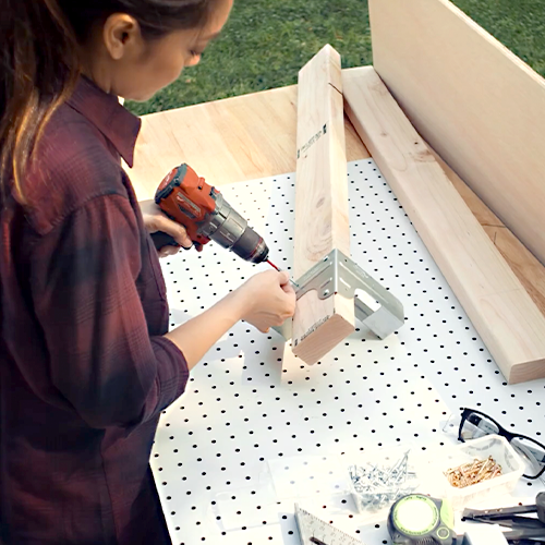 A person (Jen Woodhouse) drills screws into a wood joint using Simpson Strong-Tie hardware at a DIY workstation with tools and materials in the background.