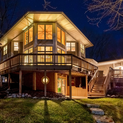 A beautifully lit modern home with an expansive wooden deck and large windows, glowing under the evening sky.