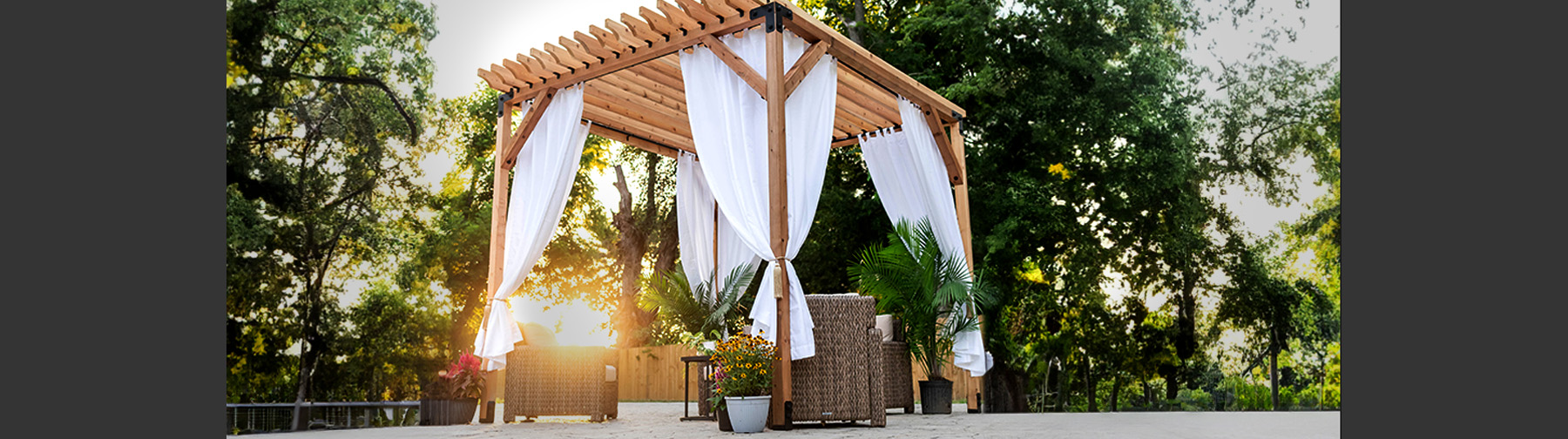 Outdoor wooden pergola with white drapes, surrounded by plants and wicker furniture, set against green trees and sunlight in the background.