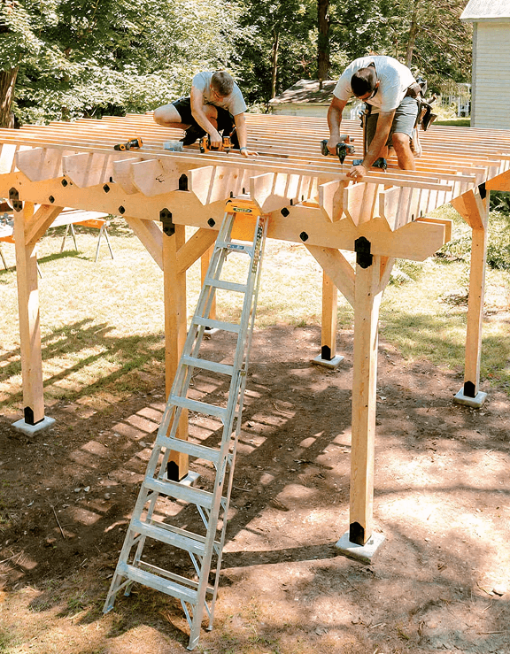 Two people build a wooden pergola frame using power tools. A ladder leans against the structure. The outdoor setting includes trees and grass in the background.