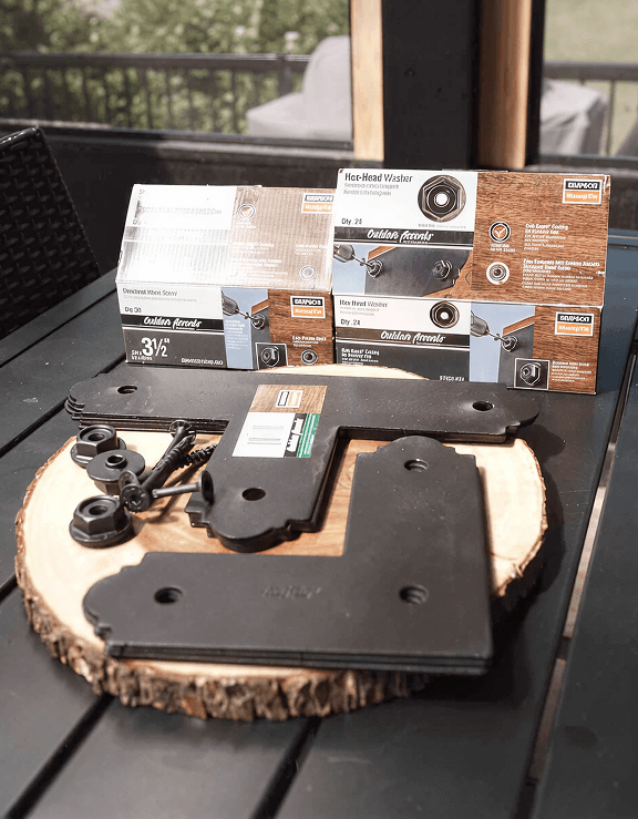 Outdoor wood screws, black metal brackets, and washers on a wooden slab. Packaging in the background reads 'Simpson Strong-Tie' and 'Outdoor Accents'.