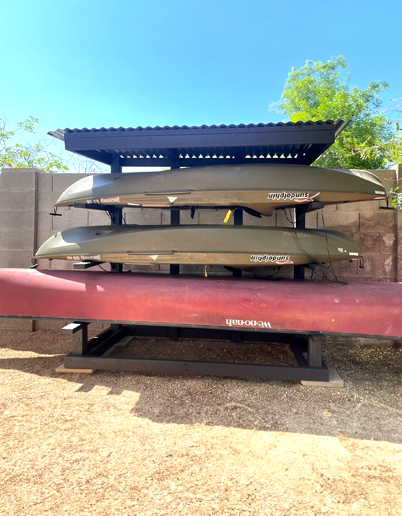 DIY outdoor rack with a corrugated roof holds multiple kayaks and a red canoe, positioned against a block wall under a clear sky.