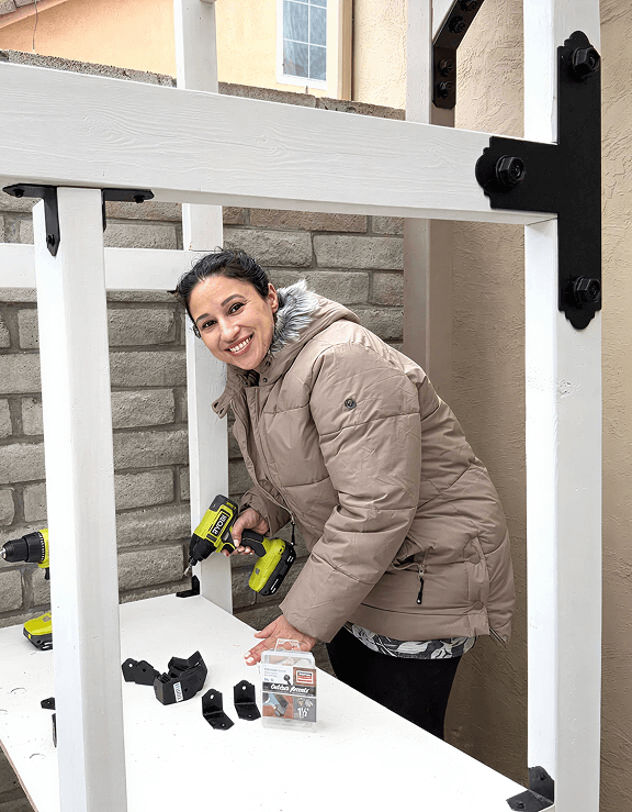 erson in beige coat uses a Ryobi drill to assemble a white wooden frame with black brackets. Tools and hardware are spread out on the table.