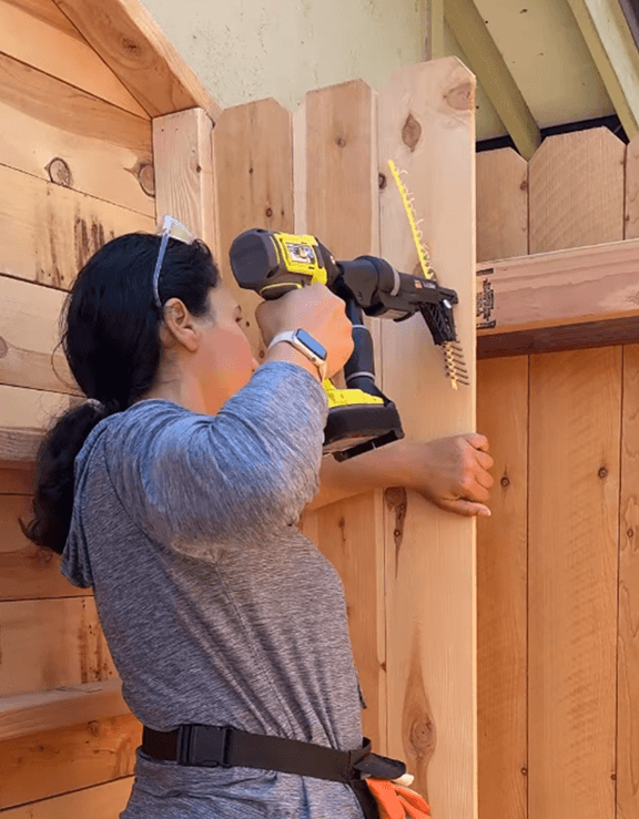 Person using a yellow power drill to attach vertical wooden boards to a wooden structure outdoors.
