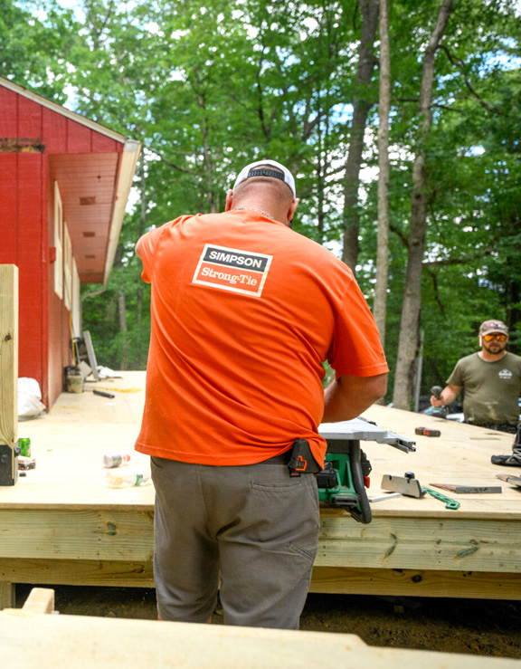 A worker in an orange Simpson Strong-Tie shirt is building a wooden deck with power tools. Another worker stands nearby, with trees and a red building in the background.