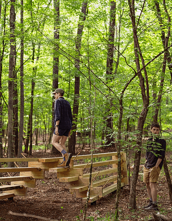 Person walking up a wooden zigzag bridge in a forest with tall green trees and another person standing nearby.