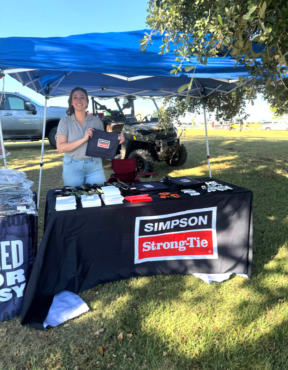 A woman standing behind a Simpson Strong-Tie promotional booth outdoors under a blue canopy, displaying branded materials and giveaways.