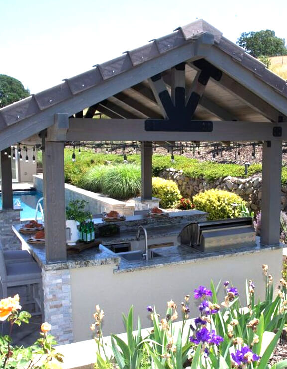 Outdoor kitchen setup with a granite countertop and wooden pergola, surrounded by vibrant flowers and greenery, showcasing a relaxing and elegant backyard space.