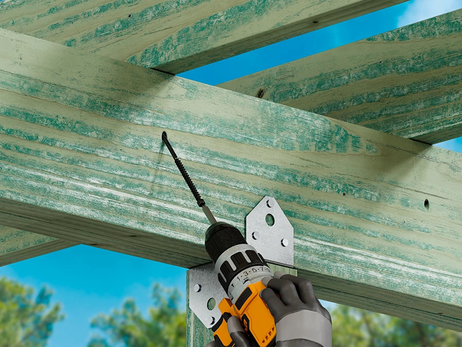 Close-up of a worker using a power drill to fasten screws into a metal connector attached to a green wooden beam structure.