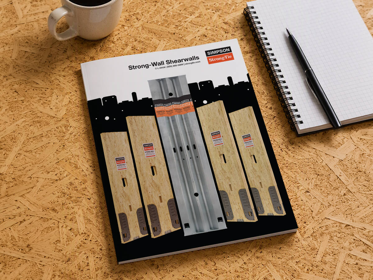 Simpson Strong‑Tie “Strong‑Wall Shearwalls” product catalog on a wood desk, a notebook, and a coffee mug.