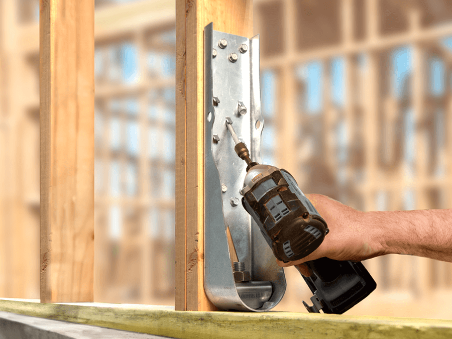 A close-up of a worker using a power drill to install a Simpson Strong-Tie HDUE Holdown on a wooden frame for added structural stability in a construction site.