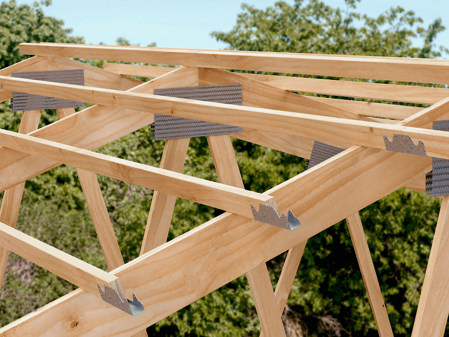Wooden roof trusses with metal connectors form a triangular pattern, set against green trees and a blue sky, showing structural roof support.