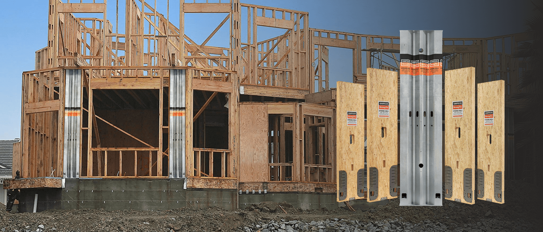 Large timber-framed building under construction at sunset, with exposed roof trusses and wood framing. In the foreground, galvanized metal post base connectors and a concrete foundation mockup with wood posts and metal brackets are displayed against the construction site background.