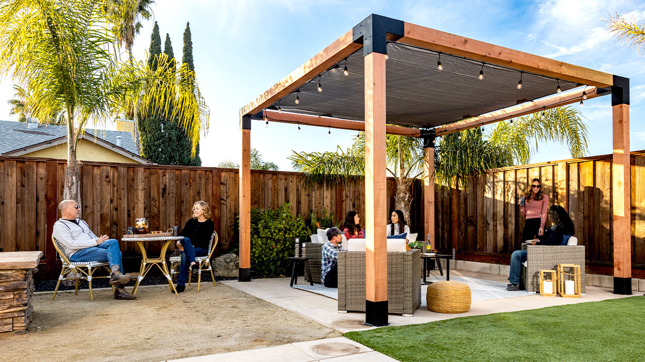 Outdoor pergola with wooden beams and a shade cover, surrounded by palm trees. People are sitting and chatting in a cozy backyard setting.