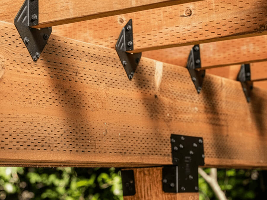 Close-up of outdoor wooden pergola beams with black metal brackets and fasteners, showing structural hardware attached to natural wood.