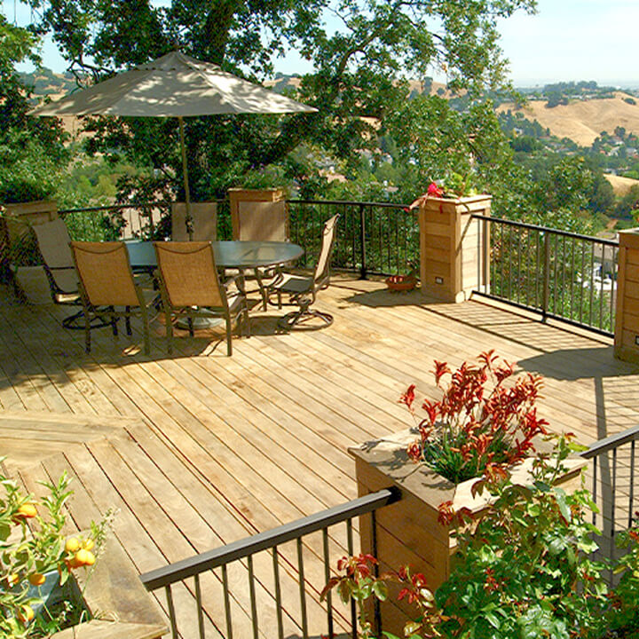 A wooden hillside deck with outdoor dining furniture, an umbrella, and planters overlooking scenic rolling hills and a valley view.