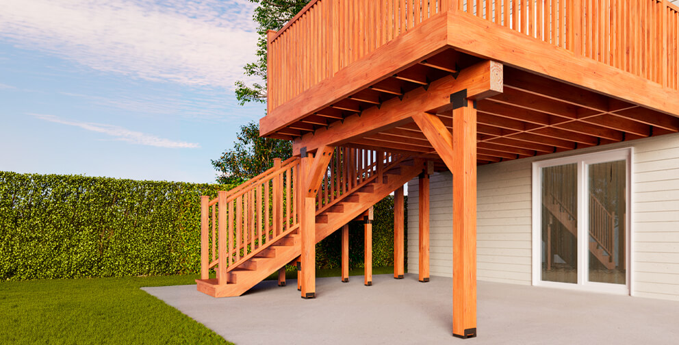 Elevated wooden deck with railing and stairs attached to a house, supported by posts over a concrete patio, with green lawn, hedge, and blue sky in the background.