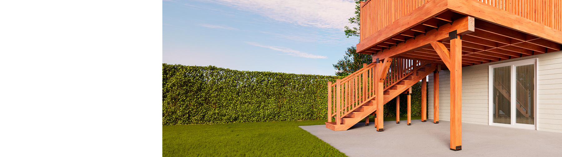 Elevated wooden deck with railing and stairs attached to a house, supported by posts over a concrete patio, with green lawn, hedge, and blue sky in the background.