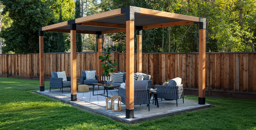 A modern wooden pergola built using the Sage System with outdoor lounge furniture on a stone patio, surrounded by a neatly manicured lawn and a fenced backyard.