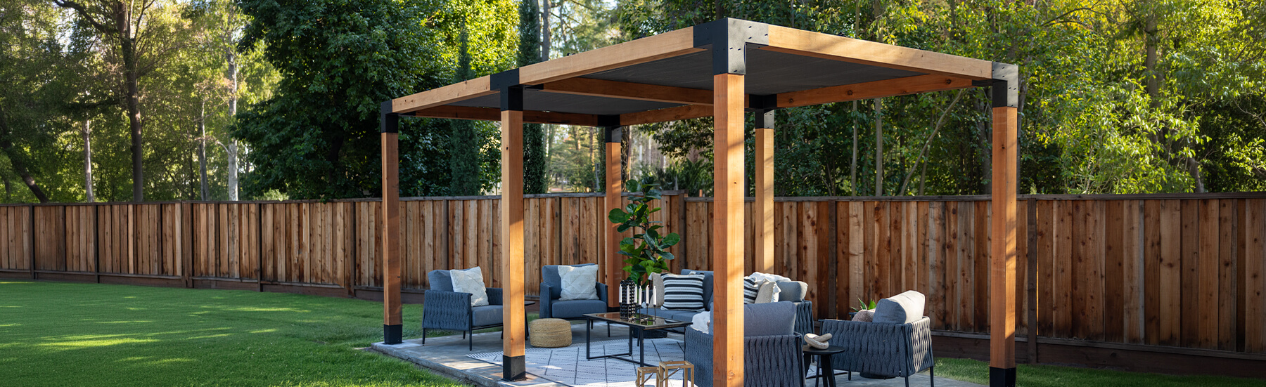 A modern wooden pergola built using the Sage System with outdoor lounge furniture on a stone patio, surrounded by a neatly manicured lawn and a fenced backyard.