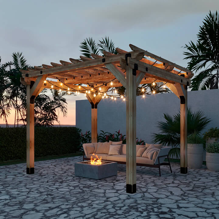 A wooden pergola with string lights illuminating a stone patio, featuring an outdoor sofa around a small fire pit at dusk, with palm plants and a garden wall in the background.
