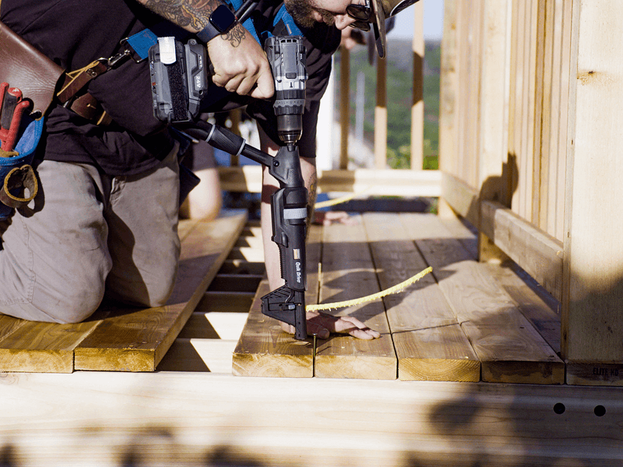 Construction worker uses a drill with a guide tool to fasten deck boards, kneeling on a wooden structure with a tape measure nearby.