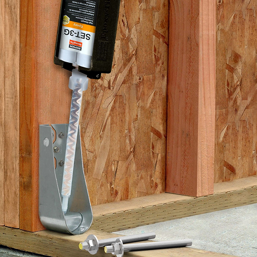 Construction adhesive cartridge dispensing into a steel anchor bracket mounted on a wood panel wall; mixing nozzle visible, with threaded anchor rods resting on the concrete base.