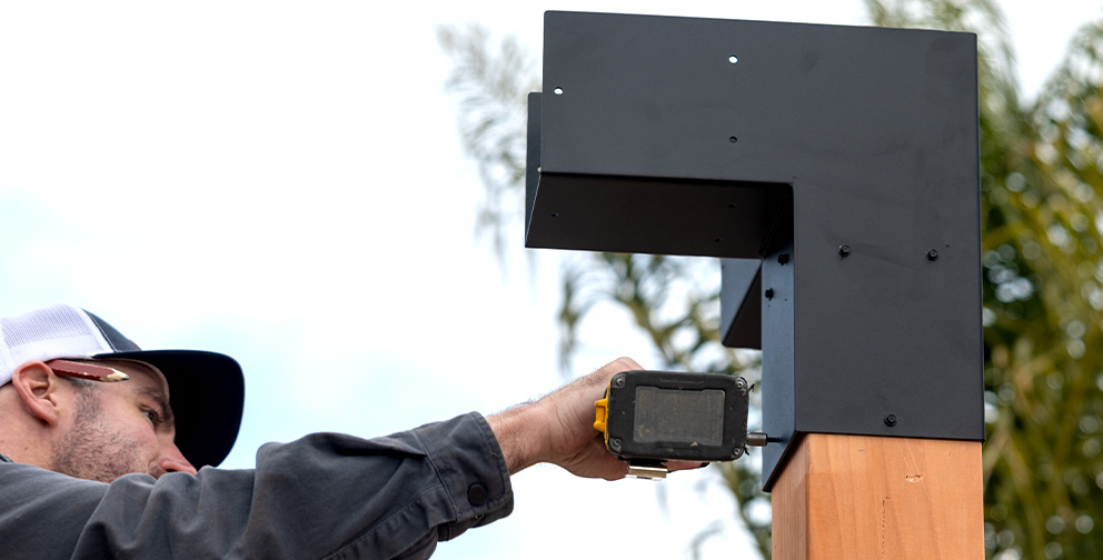 A construction worker in a black jacket and white cap uses a power drill to secure a black steel bracket onto a wooden post outdoors, with blurred palm trees in the background.