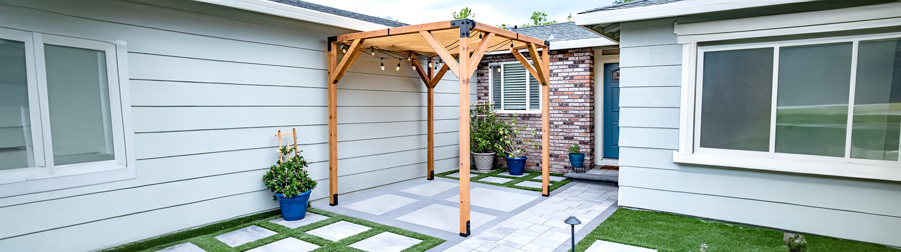 Wooden pergola installed in a small patio area between two houses, with potted plants and string lights.