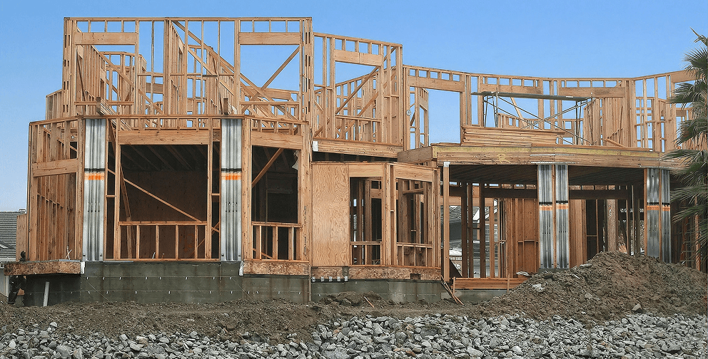 Wood-framed residential structure under construction with elevated foundation supports, exposed wall framing, and building materials visible in the foreground.