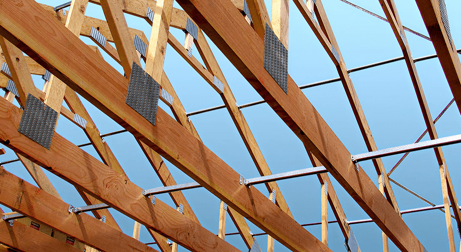Wood roof trusses with metal connector plates and bracing against a clear blue sky, showcasing structural framing in progress for a building under construction.