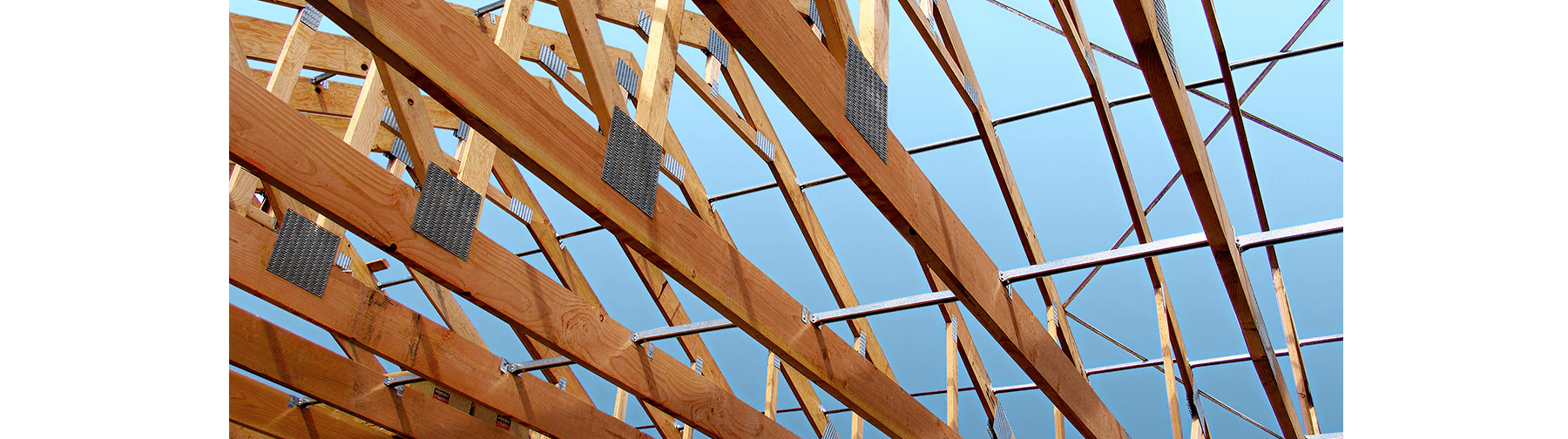 Wood roof trusses with metal connector plates and bracing against a clear blue sky, showcasing structural framing in progress for a building under construction.