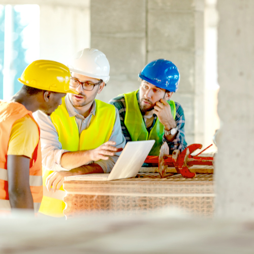 Three construction workers wearing safety gear, including hard hats and high-visibility vests, discussing plans while looking at a laptop on a construction site.