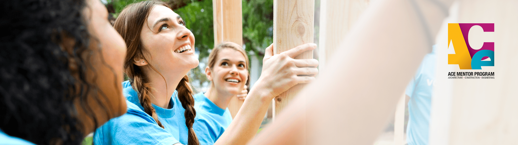 Three young women in blue shirts, smiling and working together to build a structure, with the ACE Mentor Program logo in the background.