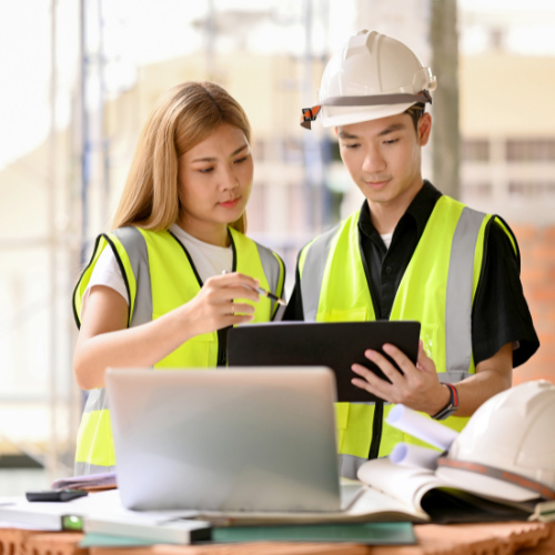 Two construction professionals wearing safety vests and helmets reviewing plans on a tablet at a construction site, with various tools and a laptop on the table in front of them.