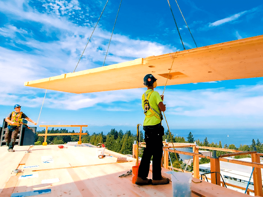 Construction crew guides a large mass timber panel into place on a multi-story building site overlooking trees and water.
