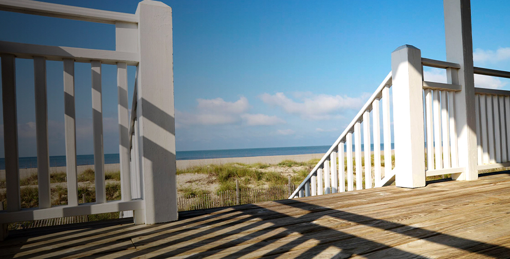 A beachside wooden deck with white railings and posts overlooks sand dunes and the ocean under a bright blue sky.