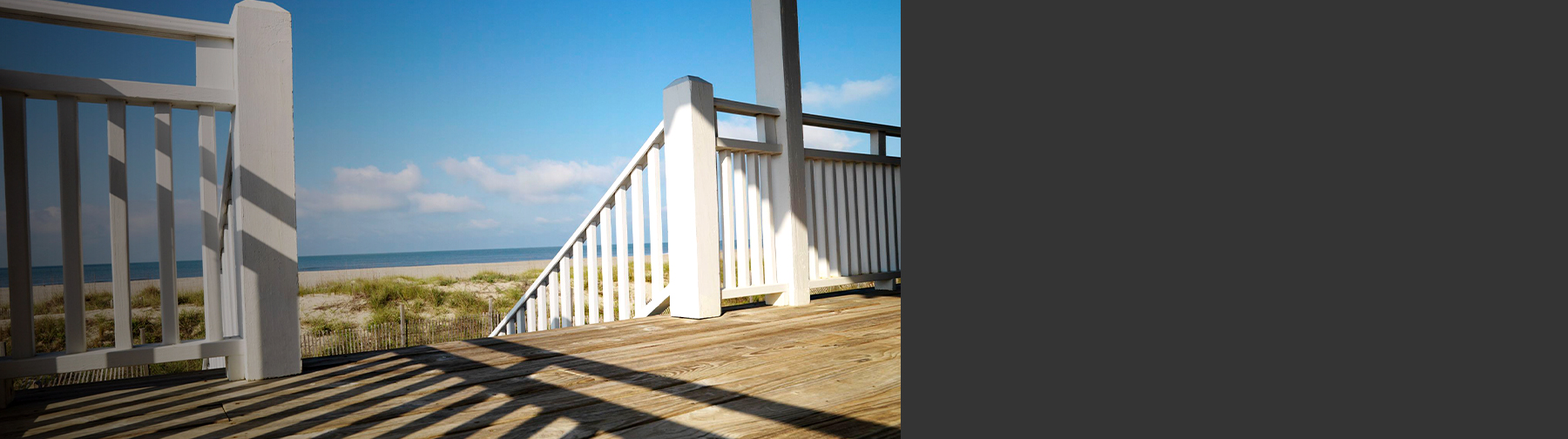 A beachside wooden deck with white railings and posts overlooks sand dunes and the ocean under a bright blue sky.