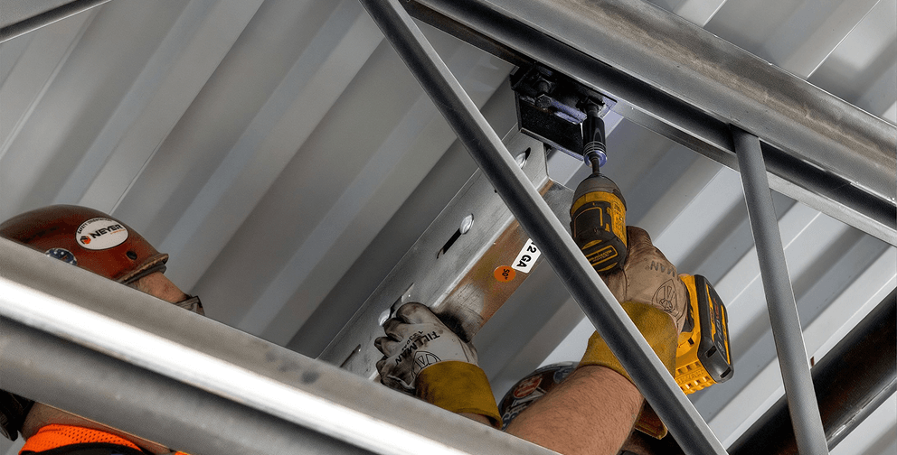 Installer secures a QuickFrames™ modular support system to structural steel using a power tool, demonstrating efficient overhead installation for electrical, mechanical, and cable management infrastructure.