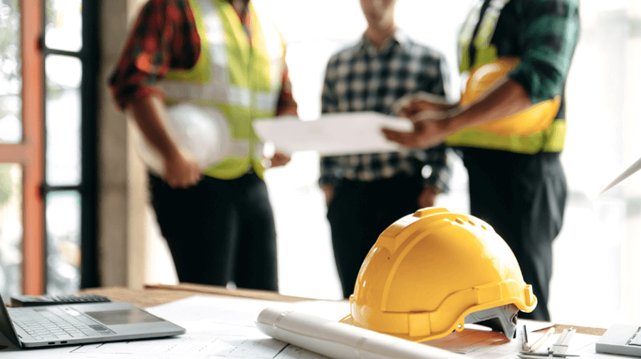 A yellow hard hat sits on a desk with construction blueprints, a laptop, and rolled-up plans, while a group of engineers and workers discuss a project in the background.