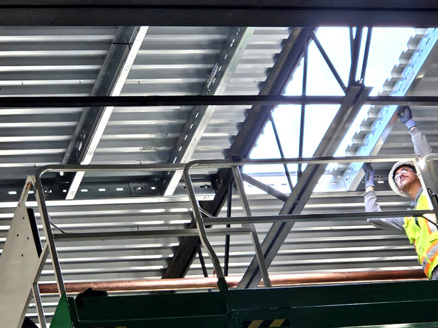 A construction worker on a lift installs a QuickFrames steel support system in a ceiling opening, ensuring structural reinforcement for rooftop equipment.