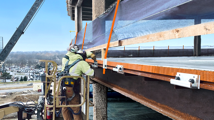 Two construction workers in safety gear securing edge tie systems to the steel structure of a building under construction. The edge tie is attached to the perimeter of the steel frame for safety and stability during construction.