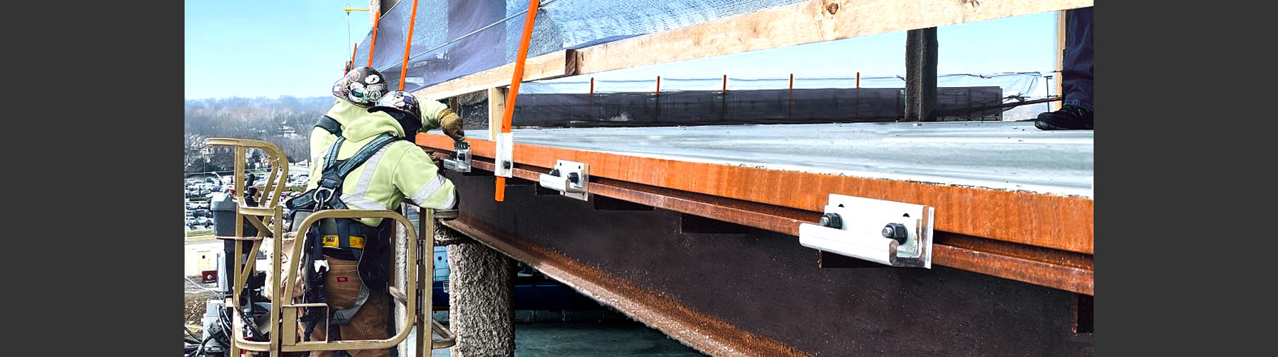 Two construction workers in safety gear securing edge tie systems to the steel structure of a building under construction. The edge tie is attached to the perimeter of the steel frame for safety and stability during construction.