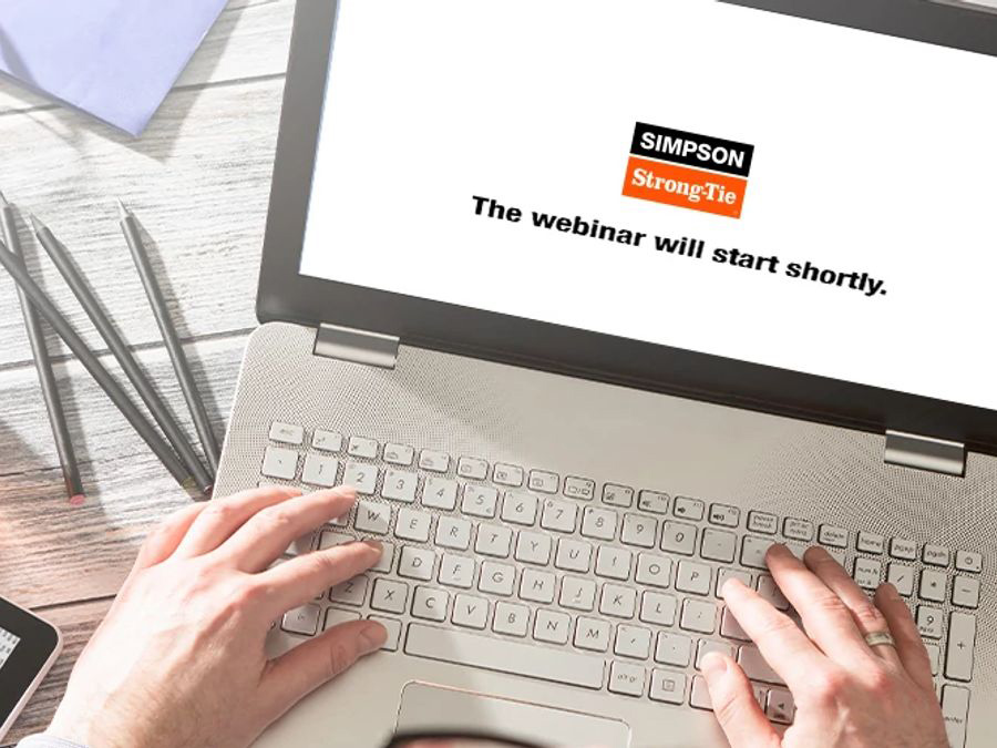 Overhead shot a hands typing on a laptop with a screen displaying logo and heading which says,"the webinar will start shortly" on a grey wooden desk with pens and a folded paper.